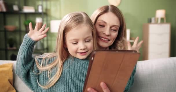 Radosna troskliwa kochająca matka siedzi z małą, słodką, białą córką na kanapie w przytulnym domu i rozmawia przez internet na czacie wideo na tablecie, machając rękami i uśmiechając się. Koncepcja dzieciństwa. Zamknij się. — Wideo stockowe