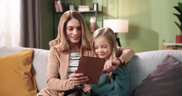 Portrait von glücklich lächelnden kaukasischen schöne Mutter verbringt Zeit mit ihrer kleinen süßen Tochter Mädchen tippen und suchen Internet auf Tablet-Gerät zusammen auf dem Sofa zu Hause sitzen. Familienkonzept. — Stockvideo