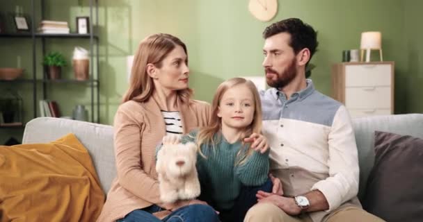 Πορτρέτο του Καυκάσου χαρούμενα χαριτωμένο γονείς ωραία οικογένεια με λίγο αξιολάτρευτο παιδί κόρη κάθεται στον καναπέ στο άνετο δωμάτιο στο σπίτι μαζί. Η μαμά και ο μπαμπάς με το μικρό παιδί κοιτάζουν την κάμερα. Έννοια ελεύθερου χρόνου — Αρχείο Βίντεο