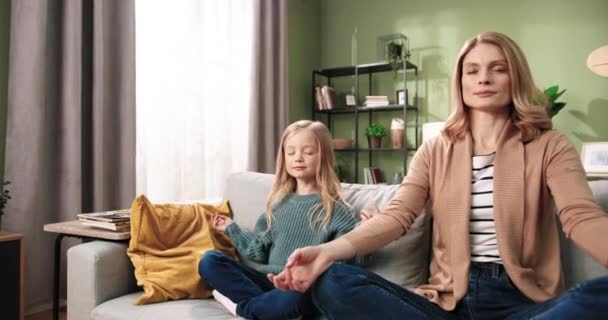 Caucasien belle famille jeune jolie mère et petite fille adorable enfant assis sur le canapé pratiquant le yoga, relaxant, méditant avec des visages heureux calmes et les yeux fermés dans une maison confortable. Santé concept de vie — Video