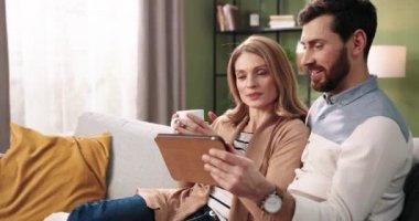 Kafkasyalı mutlu çiftin portresi internetten internetten alışveriş yapıyor tablet cihazıyla koltukta oturmuş çay içerek vakit geçiriyor. Karı koca evde dinleniyor.