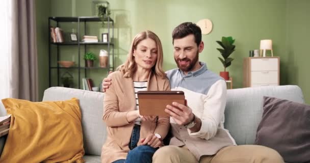 Портрет веселой молодой белой супружеской пары, печатающей и ищущей в интернете на планшете, сидящей на удобном диване, обнимающейся вместе. Семья проводит время дома с помощью гаджета — стоковое видео