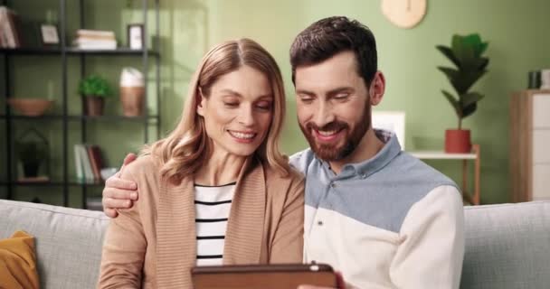 Крупным планом портрет счастливой улыбающейся белой супружеской пары супругов, ищущей в Интернете, просматривающей онлайн выбор и покупающей вещи вместе с помощью планшета, сидя на диване дома — стоковое видео