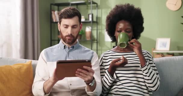 Mezcla de razas feliz pareja casada agradable sentado en el sofá descansando en casa navegando en línea en la tableta y sorbiendo bebida caliente. Concepto de comercio electrónico. Hombre escribiendo en el dispositivo y hablando con la mujer — Vídeos de Stock