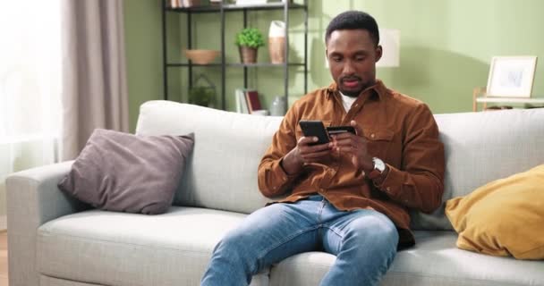 Χαρούμενος νεαρός Αφρικής Αμερικανός όμορφος άντρας κάθεται στον καναπέ στο άνετο σαλόνι στο σπίτι και περιήγηση online ψώνια στο διαδίκτυο αγορά online κάνουν την αγορά χρησιμοποιώντας smartphone και πληρώνουν με πιστωτική κάρτα — Αρχείο Βίντεο