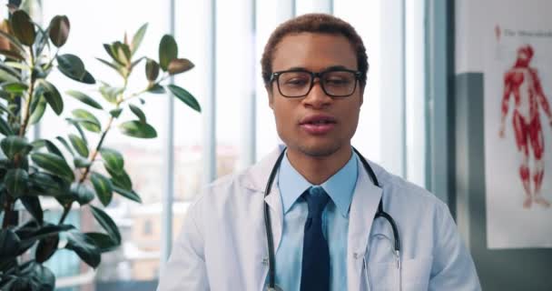 Zbliżenie przystojny radosny młody Afroamerykanin męski lekarz w biały płaszcz siedzi w szafie w szpital w miejscu pracy rozmawiając na wideo połączenie online przez kamery z pacjentem, koncepcja kliniki — Wideo stockowe