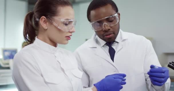 Крупный план молодых врачей женского и мужского пола в белых халатах, стоящих в клинической лаборатории в очках, делающих анализ крови, обсуждающих образец, микробиологию. Концепция анализа — стоковое видео