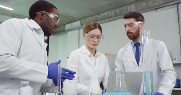 Портрет молодої команди лікарів-спеціалістів у білих халатах та окулярах, що стоять у лабораторії, що працює над ноутбуком, що розробляє вакцину із зразком, дослідженням мікробіології, медичною — стокове відео