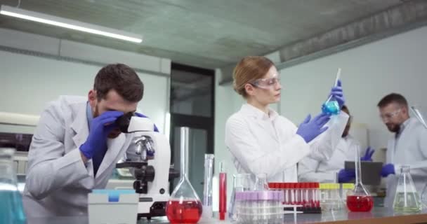 Portrait de travailleurs de laboratoire blancs, hommes et femmes, travaillant en laboratoire au microscope et au tube avec échantillon vaccinal. Des collègues de race mixte qui parlent en arrière-plan. Expérience de recherche pharmaceutique — Video