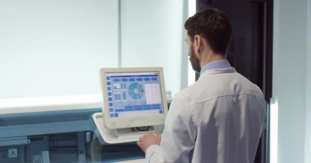 Zadní část bělošského mladého zaneprázdněného muže profesionální lékař v bílém plášti stojí v laboratoři klepání na pracovní plochu počítače, diagnostika, zkoumání vývoje vakcíny pomocí high-tech výzkumu diagnózy — Stock video