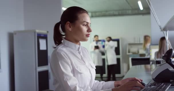 Visão lateral Retrato de caucasiano bela jovem cientista assistente de laboratório profissional se senta em laboratório moderno e digitação no teclado especialista em microbiologia do computador no local de trabalho Conceito médico — Vídeo de Stock