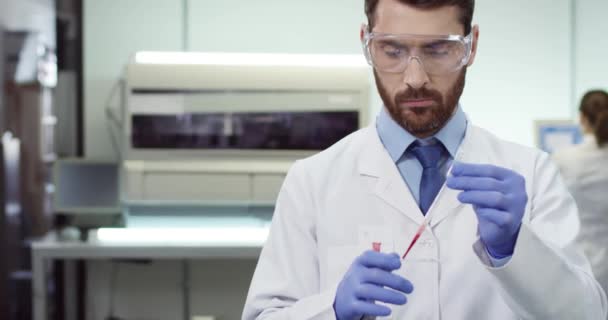 Крупный план серьезного молодого кавказского лаборанта в пальто и очках работает в современной клинической лаборатории, заливая образец крови на эксперимент по проведению стекла, концепцию анализа, генетику, микробиологию — стоковое видео