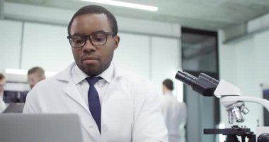 Yakışıklı Afro-Amerikan genç erkek laboratuvar uzmanının portresi hastanede mikroskop üzerinde çalışıyor ve laboratuvarda dizüstü bilgisayarda genetik araştırma yapıyor, aşı geliştiriyor.