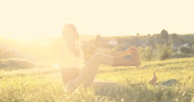 Beyaz tenli güzel bir kadın yeşil çimenlerde oturuyor ve gün ışığında yükselen ayak parmaklarını tutuyor. Sabahın erken saatlerinde oldukça sportif dişi yogiler geriniyor. Açık havada beden eğitimi..