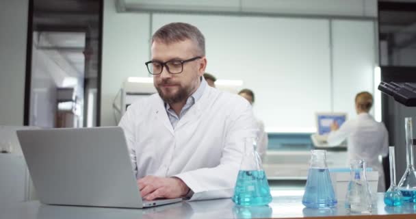 Portrait d'un médecin scientifique masculin professionnel d'âge moyen caucasien occupé naviguant en ligne sur un ordinateur portable travaillant dans un laboratoire hospitalier avec de nombreux tubes avec du liquide sur la table, développement de vaccins — Video
