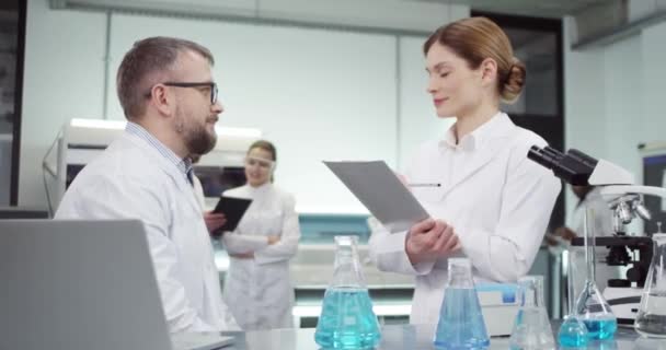 Portrait de jolie assistante médicale blanche parlant avec un médecin expérimenté d'âge moyen qui navigue sur un ordinateur portable travaillant dans une clinique avec différents tubes avec du liquide sur la table — Video