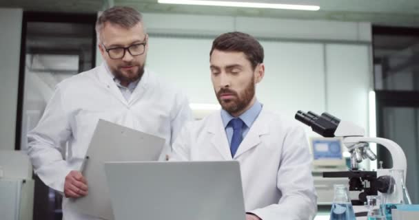 화이트 코트를 입은 바쁜 백인 남성 전문의 가인간 과학자 동료와 함께 노트북 컴퓨터로 병원 실험실에서 연구 결과와 분석 결과를 확인하는 모습. 건강 관리 개념 — 비디오