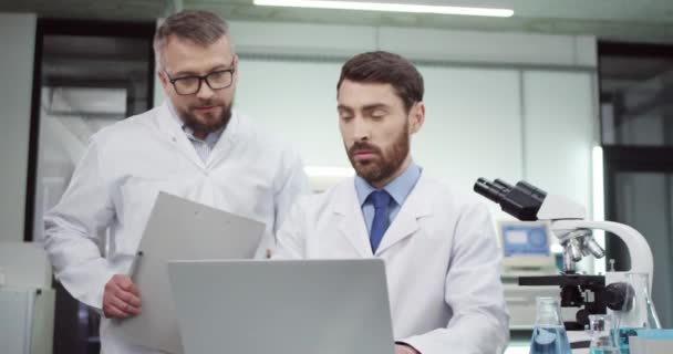 Ritratto di medico professionista caucasico impegnato in camice bianco che controlla i risultati della ricerca e l'analisi in laboratorio ospedaliero su computer portatile con collega uomo scienziato. Concetto sanitario — Video Stock