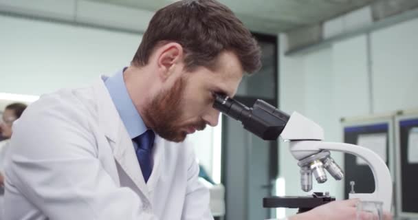 Nahaufnahme eines gutaussehenden jungen bärtigen Mannes aus dem Kaukasus, der im Krankenhauslabor mit Mikroskop arbeitet und am Laptop surft und Experimente durchführt, Forschungskonzept — Stockvideo