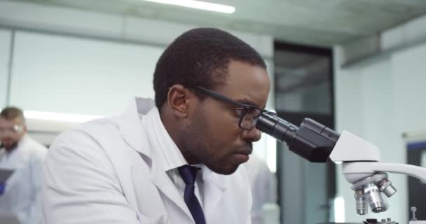 Κοντινό πλάνο του όμορφου Αφροαμερικανού νεαρού συγκεντρωμένου γιατρού, που δουλεύει στο μικροσκόπιο και στέλνει μηνύματα σε υπολογιστές laptop surfing στο εργαστήριο του νοσοκομείου. Έννοια της γενετικής έρευνας — Αρχείο Βίντεο