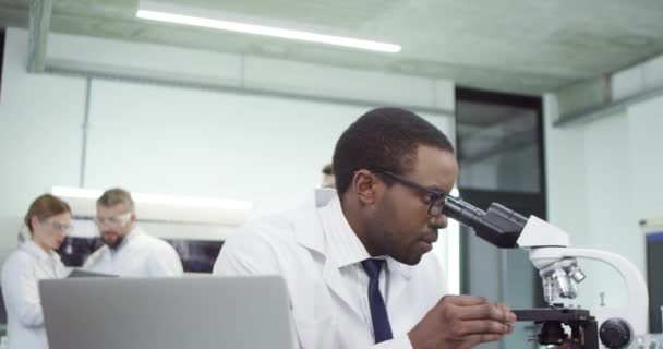 Крупным планом несчастного недовольного афроамериканского молодого занятого лаборанта работает в больнице на микроскопе и пишет на ноутбуке в лаборатории, проводя генетические исследования, разрабатывая вакцину — стоковое видео