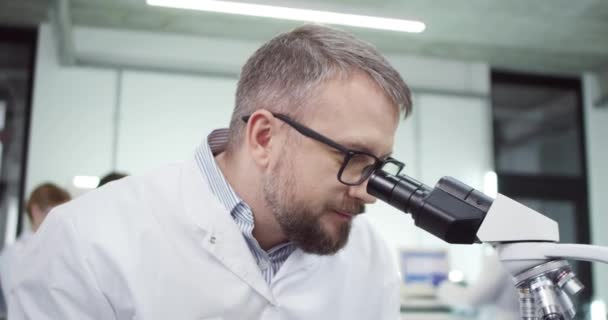 Ritratto ravvicinato del medico maschio caucasico barbuto di mezza età in occhiali che lavora in laboratorio su apparecchiature per microscopio che conducono esperimenti di sviluppo del vaccino covid-19 e digita sul computer — Video Stock