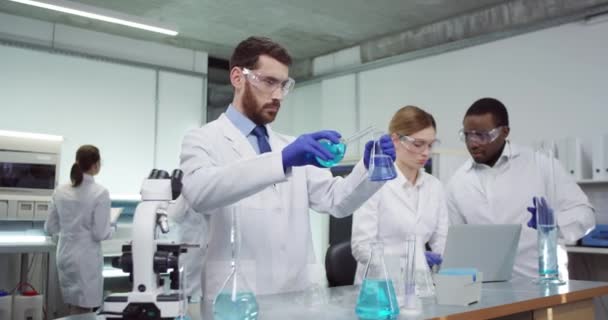 Портрет молодого кавказского бородатого ученого-микробиолога стоит в лаборатории в больнице, заливая химические жидкости в трубки, разрабатывая лекарство, в то время как сотрудники смешанных рас разговаривают и просматривают ноутбуки — стоковое видео