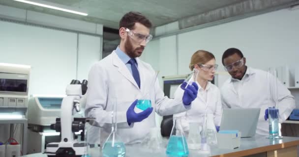 Retrato de trabajadores de laboratorio multiétnicos que trabajan navegando en la computadora portátil y hablando. Científico de laboratorio masculino caucásico mezclando químicos líquidos en vasos de precipitados llevando a cabo experimento, investigación farmacéutica — Vídeos de Stock