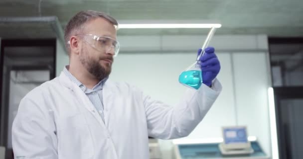 Retrato do cientista de microbiologia masculina barbudo caucasiano que trabalha na vacina em laboratório verificando o produto químico líquido azul no copo, conduzindo o experimento, conceito da pesquisa farmacêutica — Vídeo de Stock