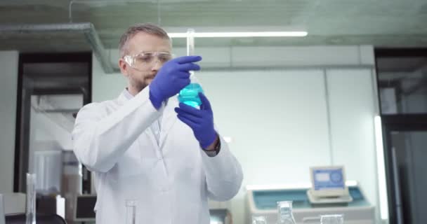 Κάμερα πλησιάζει χαρούμενη Καυκάσιος γενειοφόρος επιστήμονας εργαστηρίου στέκεται στο εργαστήριο στην κλινική εργάζονται για τη θεραπεία του ιού εκμετάλλευση ποτήρι ζέσεως με μπλε υγρό χημικό, διεξαγωγή πειράματος μικροβιολογίας έρευνα — Αρχείο Βίντεο