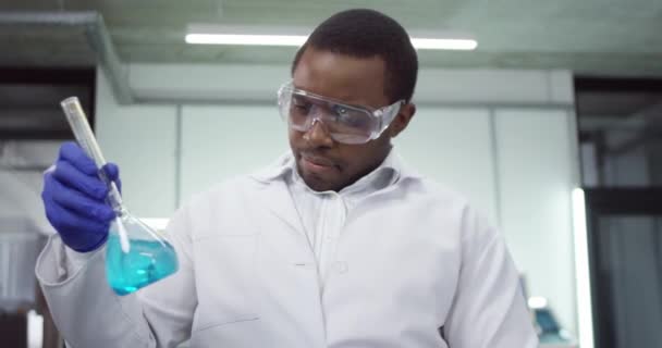 Afrika kökenli Amerikalı genç erkek mikrobiyoloji bilimadamı laboratuvarda aşılar üzerinde çalışıyor deney tüplerindeki mavi sıvı kimyasalları kontrol ediyor, tıbbi araştırmalar yapıyor. — Stok video