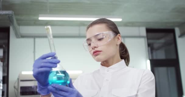 Primo piano di gioiosa bella caucasica giovane scienziata di laboratorio donna in occhiali che lavorano sul vaccino covid-19 in laboratorio tenendo becher con sostanze chimiche liquide blu guardando concetto farmaceutico fotocamera — Video Stock