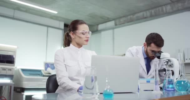 Portrait of Caucasian team of lab scientists male and female specialists working in laboratory together conducting experiment. Woman specialist typing on laptop computer while man using microscope — Stock Video