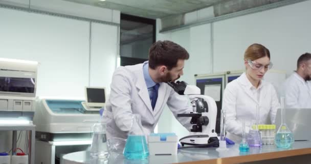 Spécialistes scientifiques masculins et féminins caucasiens occupés à taper sur un ordinateur portable et à regarder au microscope en laboratoire hospitalier parler tout en développant un vaccin. Recherche médicale, concept de microbiologie — Video