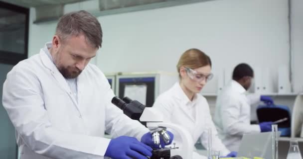 Πορτρέτο του πολυάσχολου καυκάσιου άνδρα επιστήμονα που ψάχνει στο μικροσκόπιο και μιλάει με γυναίκα γιατρό ειδικό που δακτυλογραφεί στο φορητό υπολογιστή στο εργαστήριο του νοσοκομείου Διεξαγωγή πειράματος ανάπτυξης εμβολίου covid-19 — Αρχείο Βίντεο