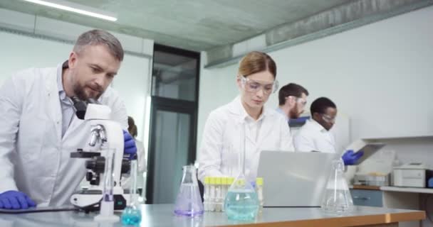 Portret zajętego zespołu medycznego kaukaskich naukowców patrzących pod mikroskopem i piszących na laptopie w szpitalnym laboratorium trzymającym zlewkę z płynną szczepionką przeprowadzającą eksperyment. Badania biotechnologiczne — Wideo stockowe
