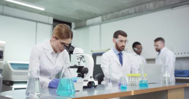 Portret poważnej białej kobiety lekarz w białym fartuchu pracujący nad mikroskopem w laboratorium rozmawiający z kolegą, który pisze na komputerze, przeprowadzający eksperymenty medyczne, badania biotechnologiczne — Wideo stockowe