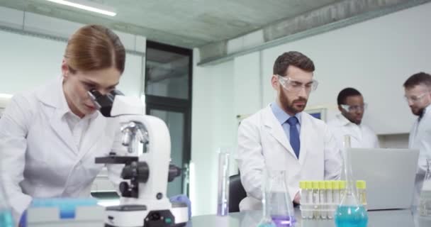Портрет занятой кавказской женщины-лаборантки в белом халате, смотрящей в микроскоп в лаборатории, разрабатывающей вакцину, разговаривающей с коллегой-мужчиной, который просматривает результаты компьютерной проверки — стоковое видео