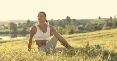 Beyaz, güzel, genç bir kadın yeşil çimlerde oturuyor ve arkasını esnemek için dönüyor. Sabahın erken saatlerinde esneme hareketleri yapan oldukça sportif dişi bir yogi. Doğa konseptinde fiziksel eğitim.