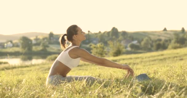 Белая красивая молодая женщина сидит на зеленой траве и наклоняется вперед к пальцам ног в солнечном свете. Довольно спортивный женский йог, растягивающий спину и ноги рано утром. Физическая подготовка. — стоковое видео