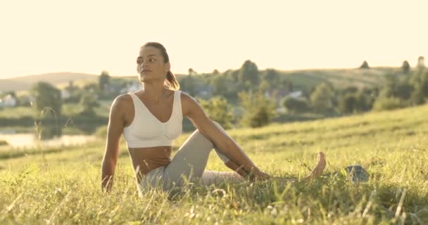 Belle jeune femme blanche assise sur de l'herbe verte et se retournant vers le côté extensible, la hanche et le dos. Plutôt sportive yogi féminin qui s'étire tôt le matin. Concept d'entraînement physique à la nature. — Video