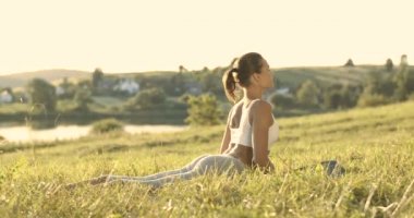 Kafkas genç dişi yogi çimlerin üzerinde yatıyor, sonra meditasyon yapmak için kobra pozuyla kalkıyor. Güzel kırsal doğa. Yoga yapan sporcu kadın, yaz sabahından itibaren erken saatlerde başlıyor. Beden eğitimi.