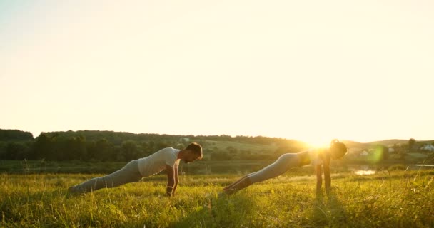 Caucasiano masculino e feminino yogis de pé na prancha na grama verde para baixo com belo sol e natureza no fundo. Homem e mulher fazendo ioga de manhã cedo. — Vídeo de Stock