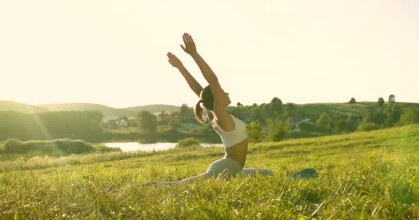 백인아름다운 젊은 여성이 푸른 잔디에 앉아 햇빛을 받으며 손을 들었습니다. 꽤나 활발 한 여성요기가아 사나에서 이른 아침에 펼쳐 집니다. 신체적 훈련. — 비디오