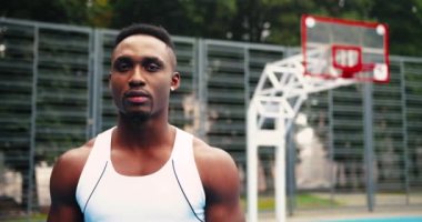 Yakışıklı Afrikalı Amerikalı basketbolcunun çocuk parkında durup kameraya bakan portresini yakından çek. Sporcu kentin açık alanındaki kortta duruyor. Beyaz adam arka planda oynuyor.