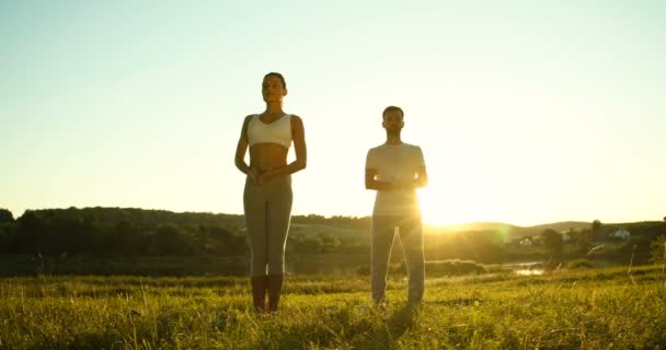 Jeune couple caucasien de yogis debout à la nature le matin dans asana et levant les mains vers le haut. Homme et femme respirant, relaxant et faisant du yoga. Journée de départ sportive masculine et féminine. Instructeur avec le gars. — Video