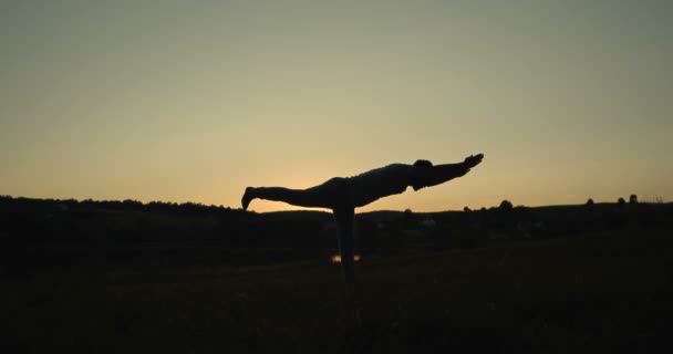 Donker mannelijk silhouet doet yoga krijger evenwicht poseren op neer buiten. Handen omhoog en één been terwijl je balanceert op de natuur. Yogi in asana in het veld als de zon opkomt vroeg in de ochtend. — Stockvideo