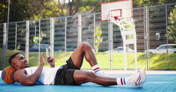 Fröhlicher afroamerikanischer männlicher Athlet, der mit dem Kopf auf dem Ball auf dem städtischen Basketballplatz liegt und draußen auf dem Handy SMS schreibt. Handsome happy man player ruhend und tippt auf dem Smartphone in der Stadt. — Stockvideo