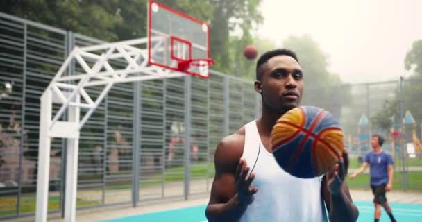 Primer plano retrato de jugador de baloncesto afroamericano guapo de pie en el patio de recreo con la pelota en las manos y mirando a la cámara. Hombre caucásico lanzando pelota en el fondo. Concepto de Streetball — Vídeo de stock