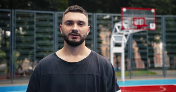 Закріпіть портрет гарного кавказького баскетболіста, що стоїть на ігровому майданчику і дивиться на камеру з спокійним обличчям. Спортсмен стоїть на вулиці міського двору в місті. Спортивна концепція — стокове відео
